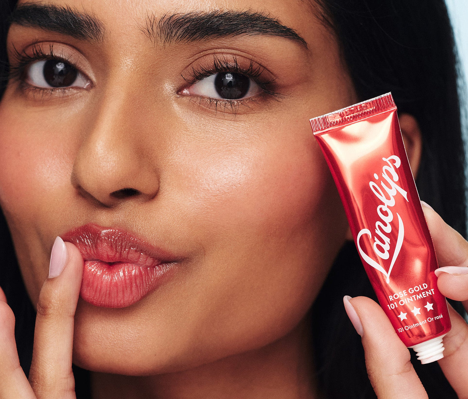 Close-up shot of a model applying Lanolips Rose Gold 101 Ointment to her lips while holding a metallic red tube near her face. This super rich balm, infused with lanolin and a subtle rose gold shimmer, provides deep hydration and a natural rosy flush for both lips and cheeks