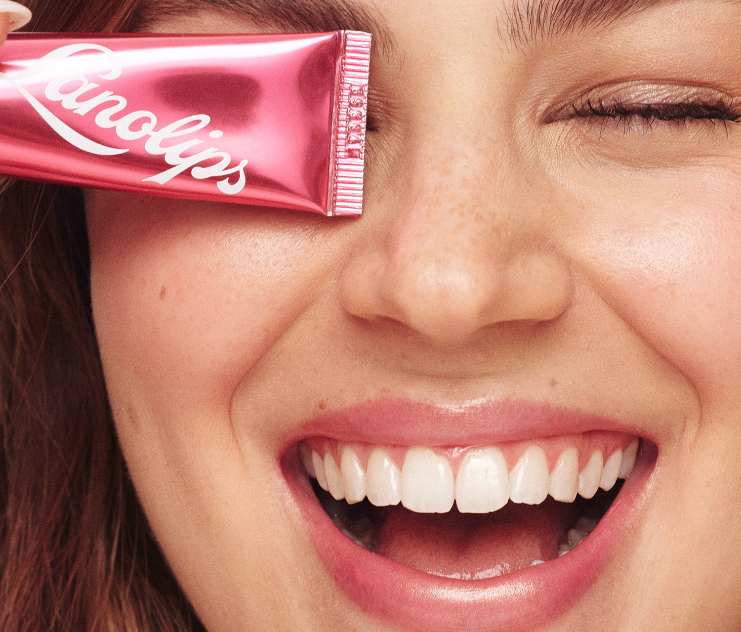 A close up shot of a model wearing the Lanolips Baby Rose Gold 101 Ointment on her lips and cheeks. The model is holding a tube, covering their left eye. their lips have a soft pink tint with a subtle sheen. Lanolips Baby Rose Gold 101 OIntment contains ultra-pure grade lanolin formula deeply moisturises while natural oils and waxes lock in hydration, leaving the lips looking plump and nourished. The model’s complexion appears luminous, with a soft golden highlight on her cheekbones.