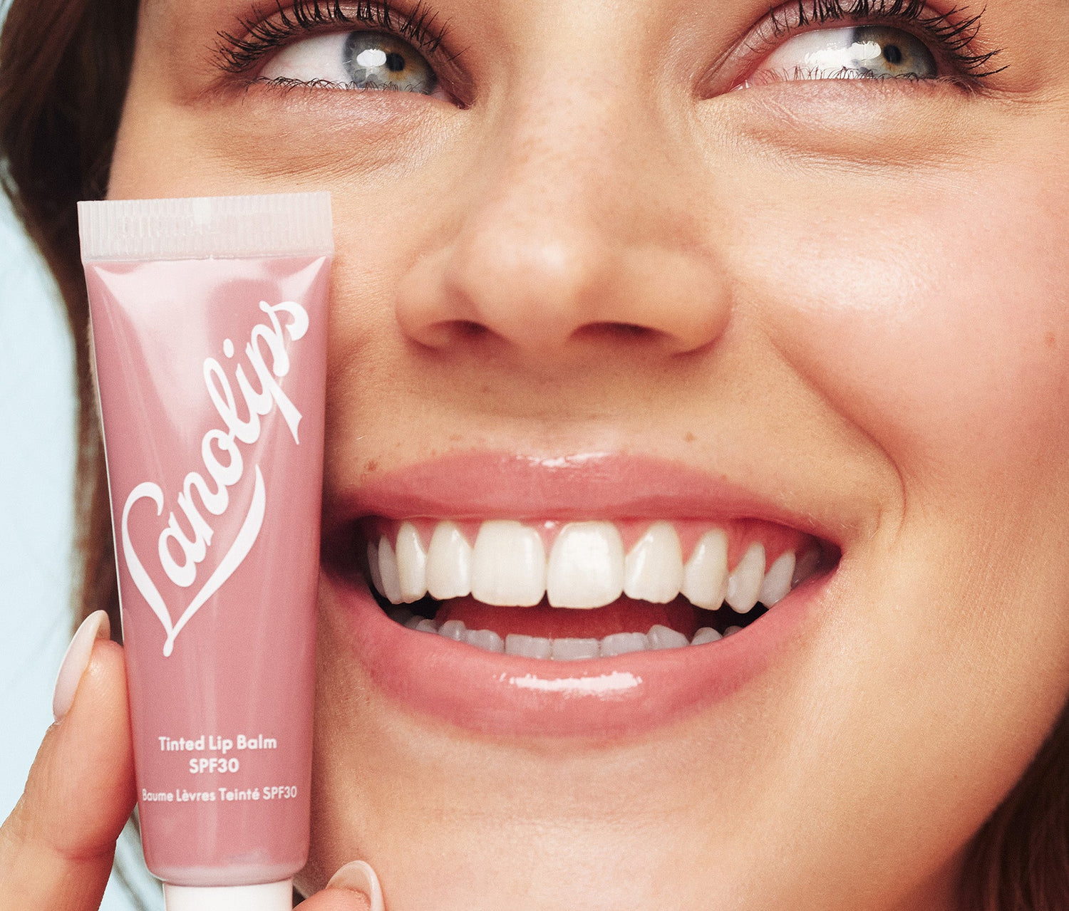 A model holding a tube of Lanolips Tinted Lip Balm SPF30 Perfect Nude close to her face. Their lips have a sheer, creamy nude tint with a soft, hydrated glow. Made with lanolin, shea butter, vitamin e and SPF30, this lanolin rich lip moisturiser deeply nourishes while protecting against sun damage. The sheer colour tint suits all skin tones, giving a natural effortless look - perfect for casual outings or weekend coffee runs.