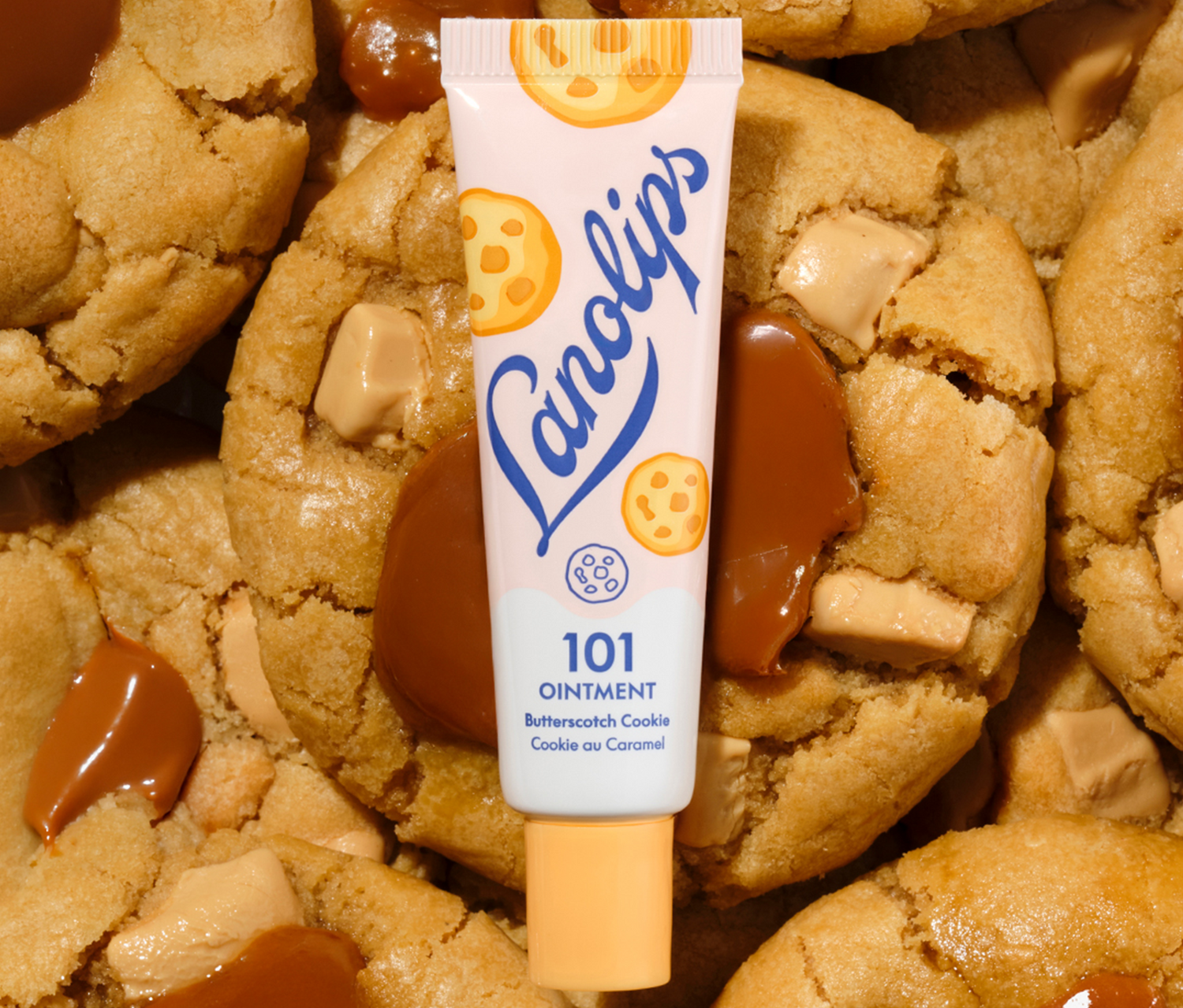 Lanolips 101 Ointment Multi-Balm in Butterscotch Cookie flavour from the 101 Delicious collection. A close-up of the 100% natural lanolin tube nestled amongst warm, golden biscuits with gooey caramel and butterscotch chunks.